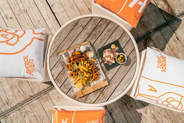 Plateau de tapas sur une table en bois entourée de coussins Aperol