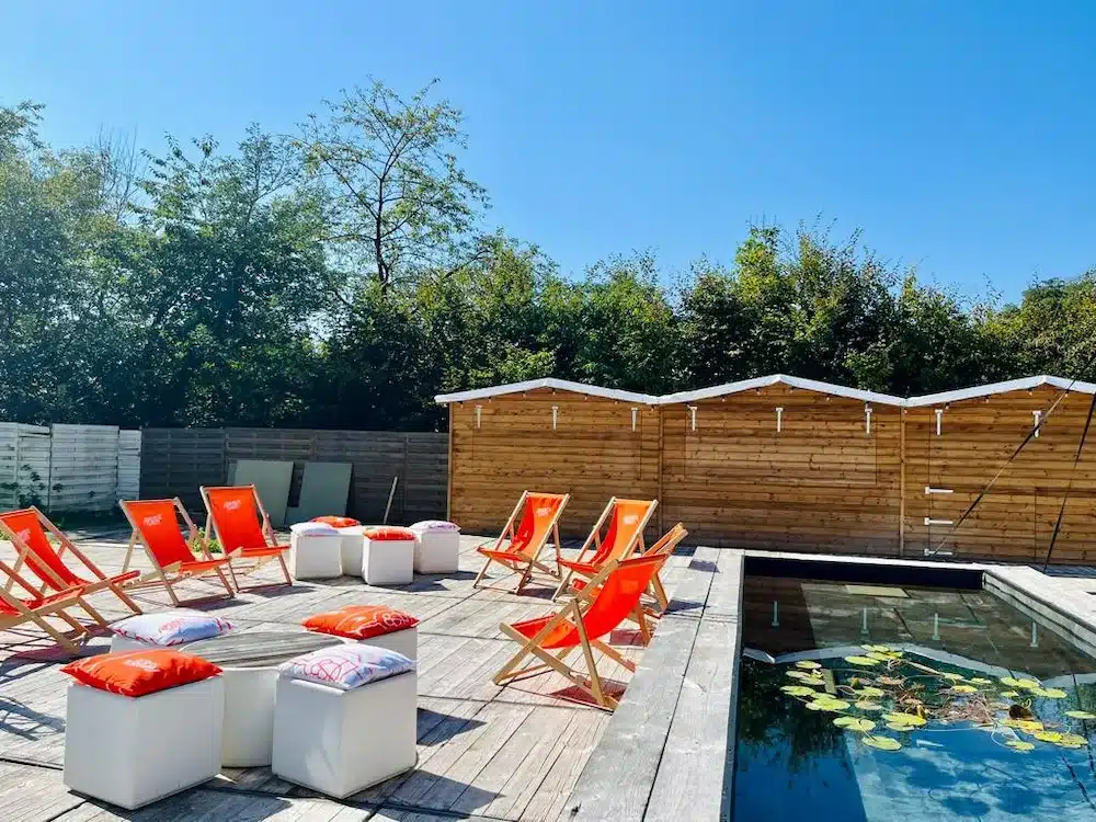 Un étang et une terrasse où se trouvent des chaises longues oranges et des poufs