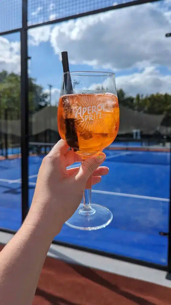 Verre d'Aperol Spritz tenu par une main devant un terrain de padel